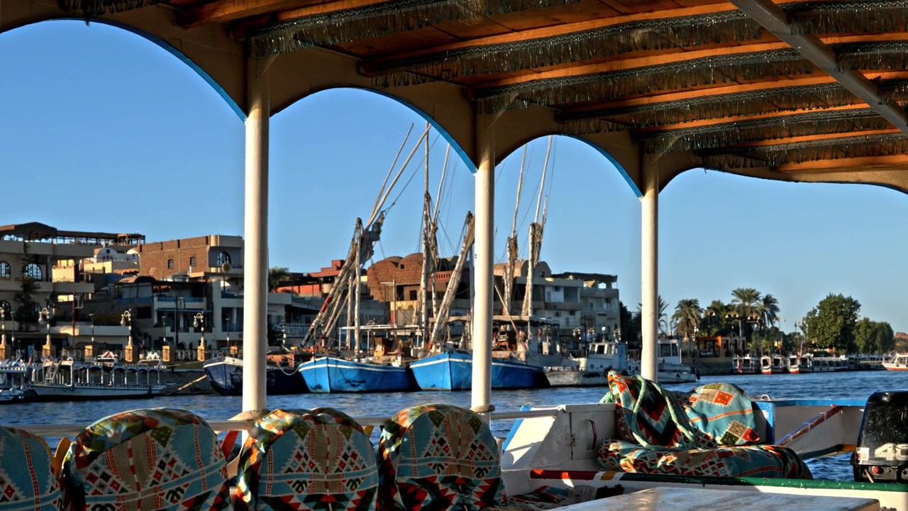 Uma vista emoldurada pela cobertura arqueada de um barco turístico com vista para o Rio Nilo em Luxor, mostrando felucas e veleiros tradicionais coloridos atracados perto da margem urbana.