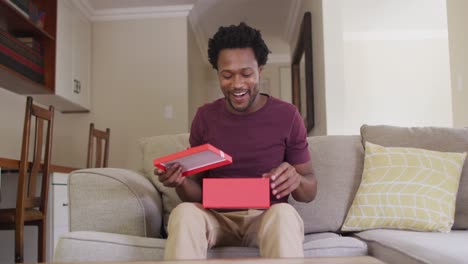 feliz hombre biracial sentado en el sofá, teniendo valentines videollamada y apertura de regalo