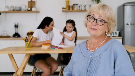 Lächelnde-ältere-Frau,-Die-Während-Eines-Videoanrufs-Zu-Hause-In-Die-Kamera-Schaut,-Während-Im-Hintergrund-Ein-Kleines-Mädchen-Und-Ihre-Mutter-Am-Tisch-Sitzen-Und-Zusammen-Zeichnen