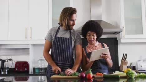 pareja de raza mixta usando delantales usando tableta digital y cortando verduras juntos en la cocina