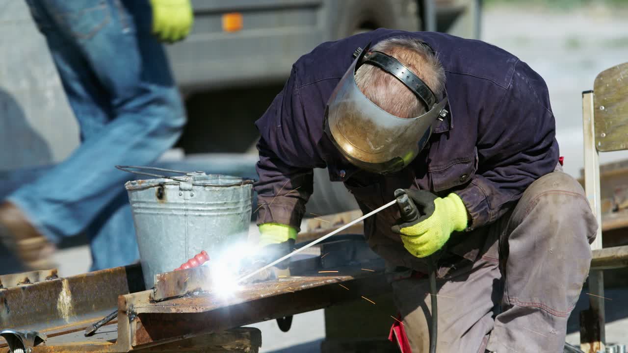 Worker in protective gear welding steel beams, harsh sunlight, slow motion