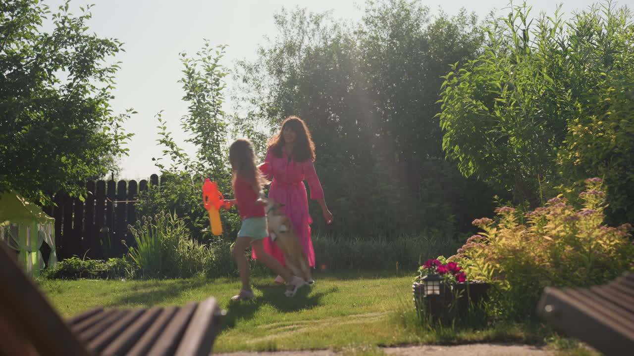 Caucasian Woman Child Playing Tag In Sunlit Garden Near Flowerbeds And Fence, Warm Summer Light, Playful Chase With Dog Darting Between Legs, Pink Dress And Bright Toy Adding Color, Cheerful Family