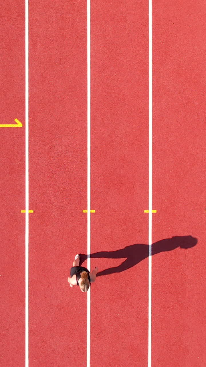 Una ripresa verticale dall'alto di una giovane donna che scatta su una pista di atletica rossa in uno stadio. Perfetto per contenuti di fitness, motivazione sportiva e allenamento atletico.