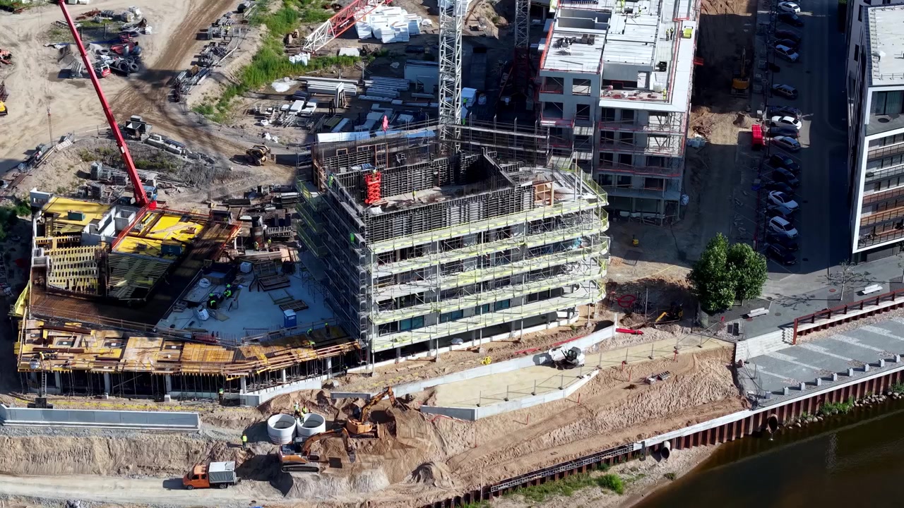 Dronevideo toont een meerverdiepingen tellend gebouw in aanbouw naast een rivier, omringd door kranen, steigers, machines en materialen. Fel daglicht benadrukt de stedelijke vooruitgang