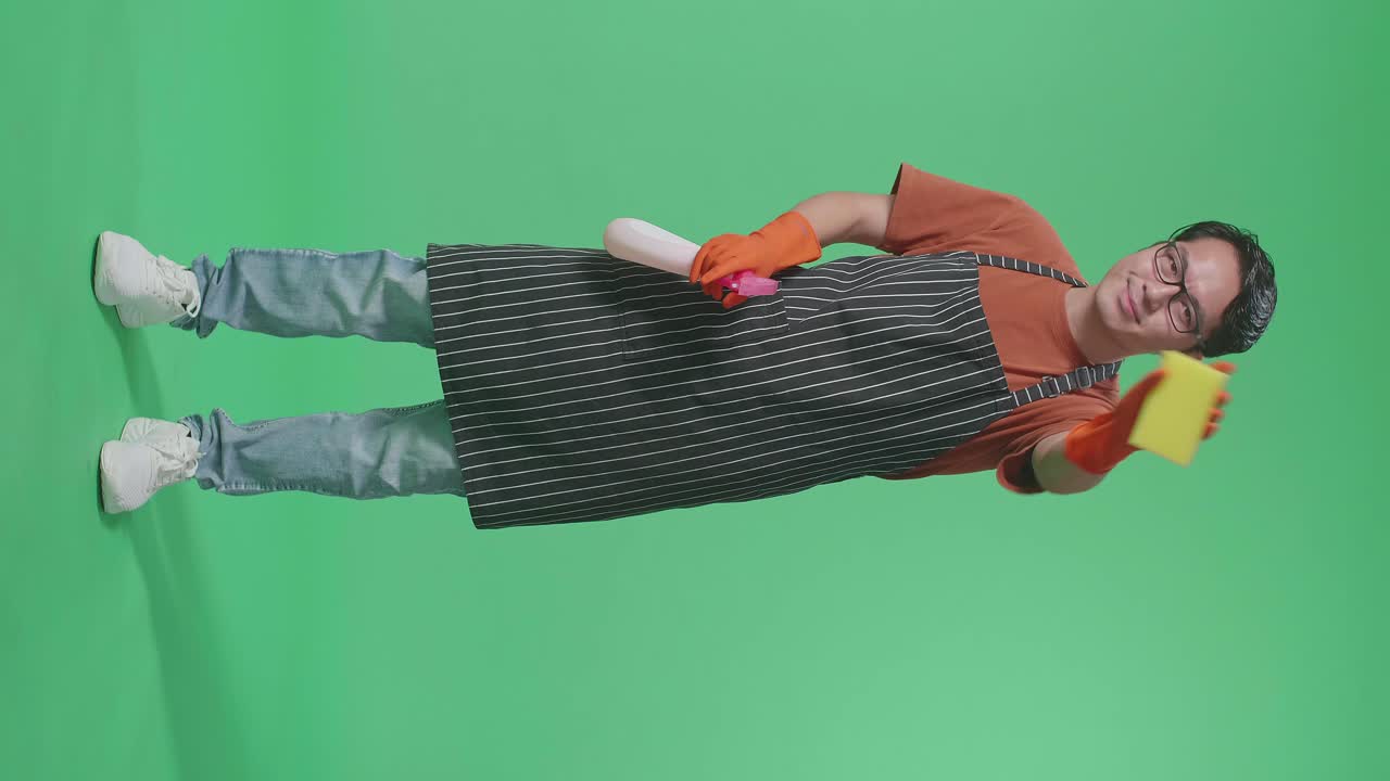 Full Body Of Asian Male Housekeeper With An Apron Washing Glass By A Spray And Kitchen Sponge In The Green Screen Studio