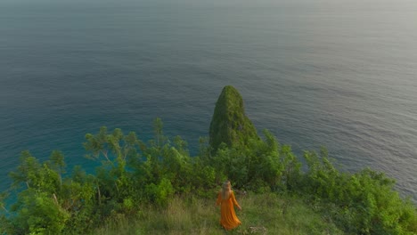 Woman-in-orange-dress-walking-at-cliff-edge-admiring-magical-sunset,-reveal-shot-of-tall-lush-rock-formation,-aerial