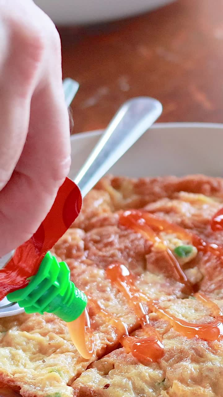 한 손은 태국 스타일의 오멜 위에 칠리 소스를 부어 맛을 높인다. 자연 조명이 있는 나무 테이블 위에 장면이 설정되어 있다.