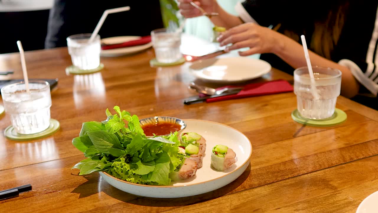 Hands preparing Vietnamese pork wraps with fresh greens on a wooden table in a well-lit restaurant setting