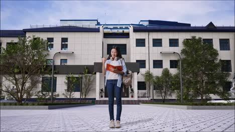 full body of asian teen girl student taking note on notebook and celebrating while standing in front of a school building