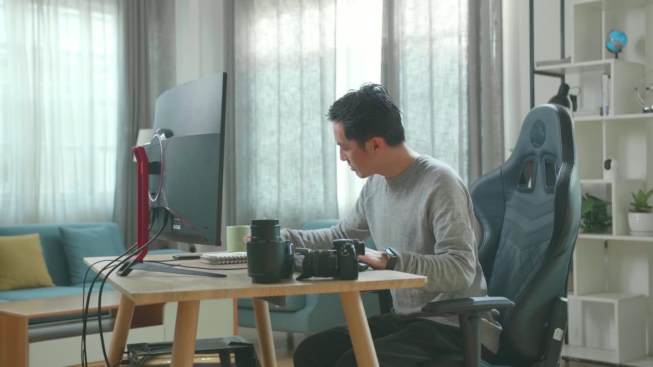 Asian Cameraman In Long Sleeved T-Shirt And Black Pants Looking At Desktop Computer Screen And Writing In Notebook On A Table While Working At Home.
