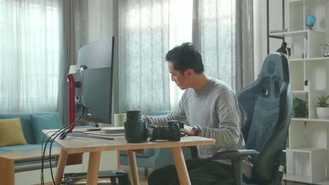 asian cameraman in long sleeved t-shirt and black pants looking at desktop computer screen and writing in notebook on a table while working at home.