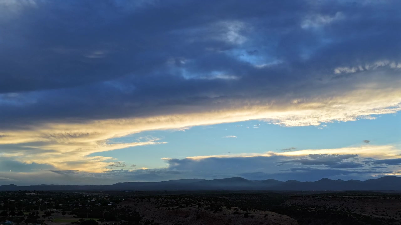 4K UHD drone timelapse capturing a striking contrast of deep blue and glowing gold sunset clouds. The vibrant sky illuminates the silhouetted mesas and distant mountain range at twilight.