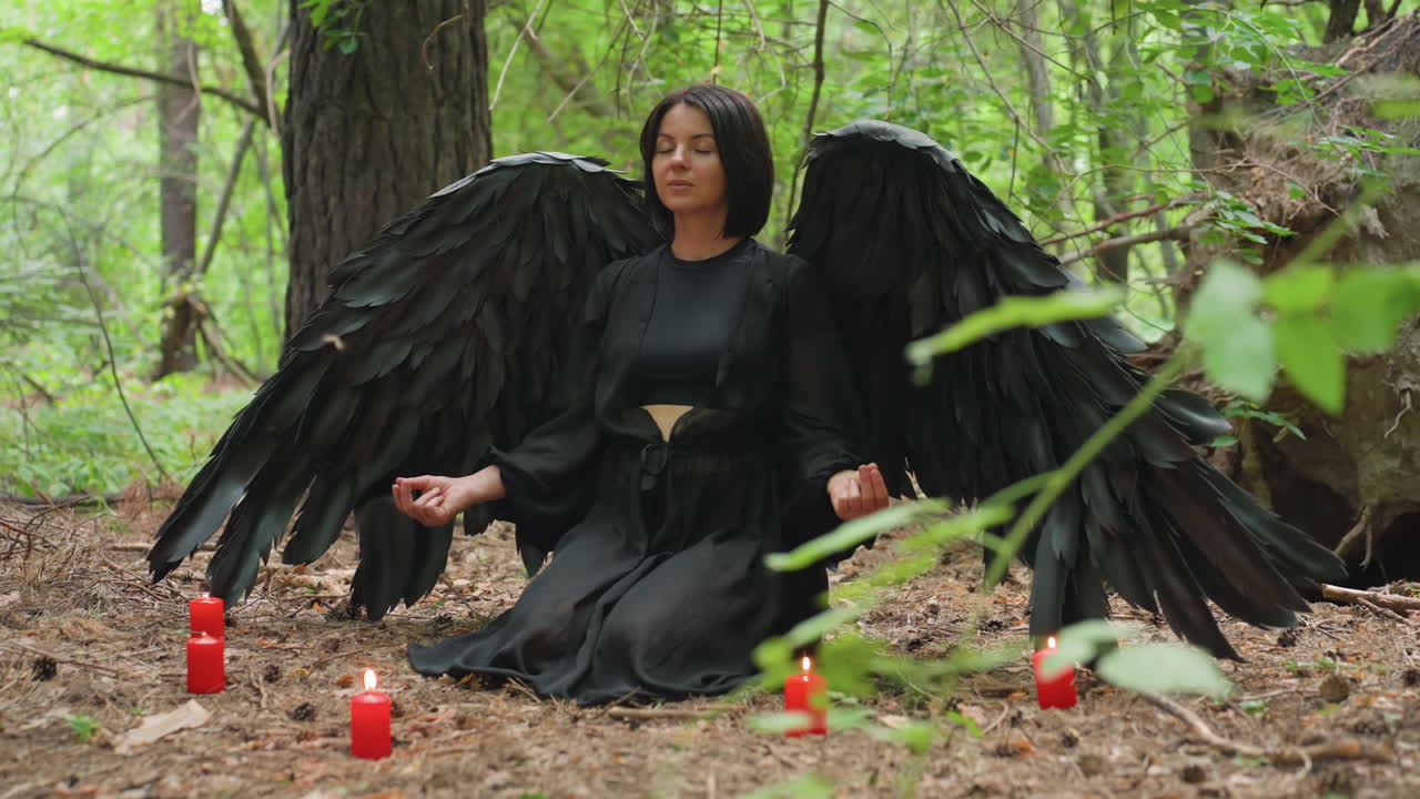 Donna con ali nere inginocchiata sul suolo di una foresta in meditazione, circondata da candele rosse incandescenti, mentre esegue un rituale pacifico in una tranquilla atmosfera boschiva piena di calma spirituale
