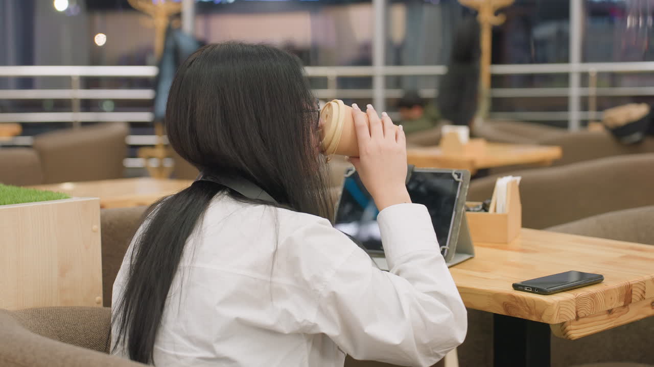 close up of woman interacting with tablet while drinking juice in cozy indoor cafe with blurred background showing other seated people and soft lighting in relaxed atmosphere