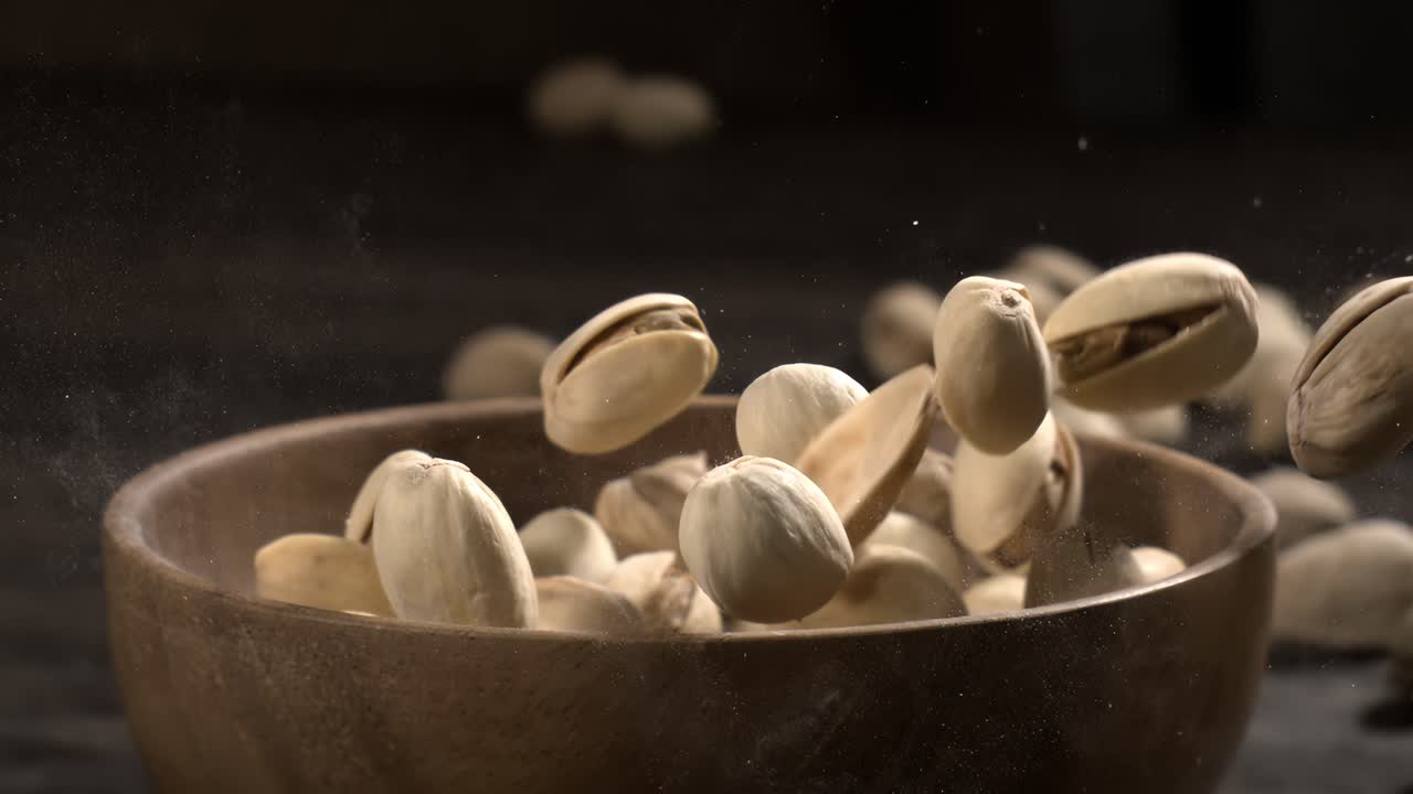 Ciotola di legno piena di pistacchi su tavolo di legno scuro, gustosa frutta secca salata sparsa intorno, concetto di snack salutare e cibo naturale. Scatto con telecamera super slow motion a 1600 fps