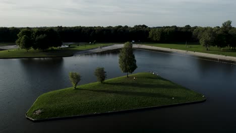 aerial rotate pan around single grass island with three trees and sunset shadow in recreational pond in the netherlands seen from the air with landscaped environment including man made beach