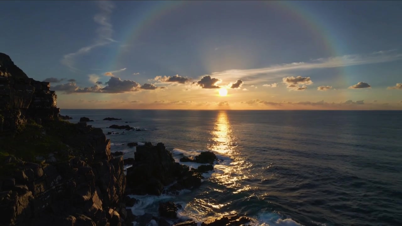 Drone siguiendo el descenso del sol sobre una costa rocosa al atardecer, filmando los cambios en el halo y los reflejos