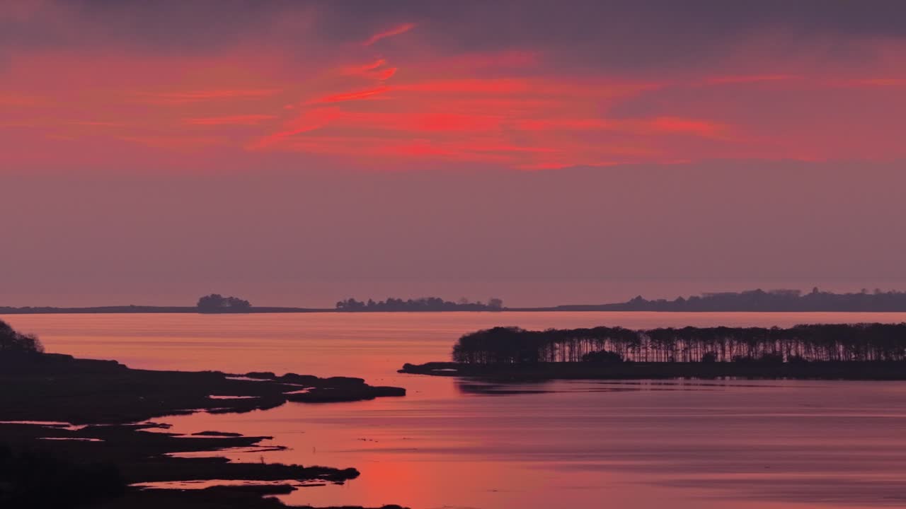 Dramatic red and orange sunset reflects on still water with dark islands and trees creating a peaceful nordic coastal mood