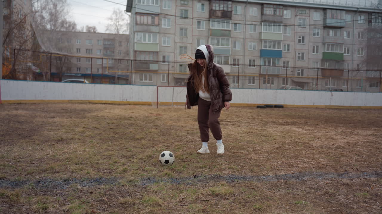 Sesión individual de regates con una mujer blanca con abrigo marrón y sudadera con capucha, control de balón en un campo embarrado, bloques de apartamentos y valla de fondo, zapatillas casual y pasos concentrados, entrenamiento independiente