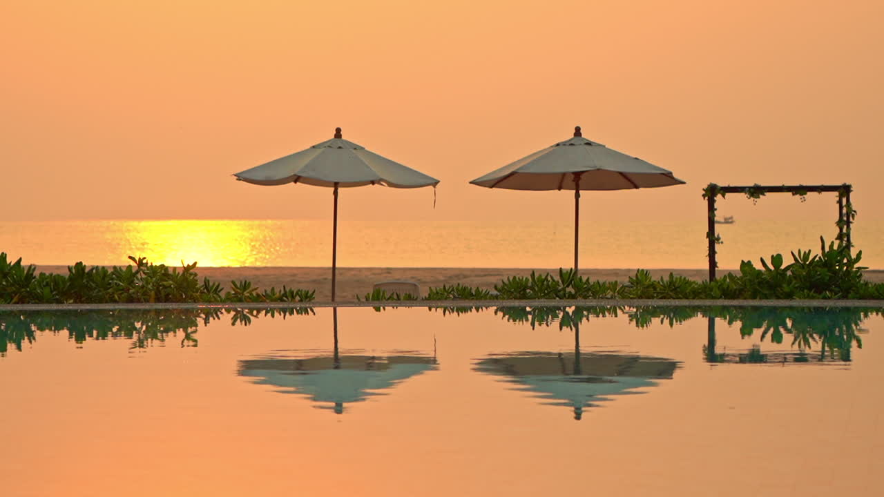 Un tramonto arancione riempie il cielo serale riflettendosi in una piscina dove due ombrelloni da spiaggia sono vuoti.