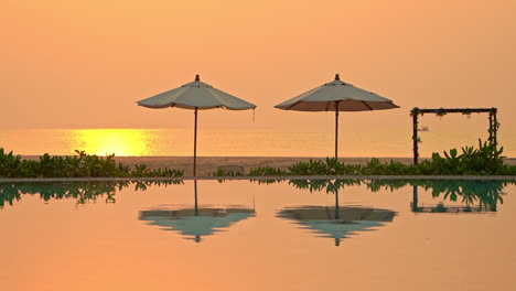 Ein-Orangefarbener-Sonnenuntergang-Füllt-Den-Abendhimmel,-Der-Sich-In-Einem-Schwimmbad-Widerspiegelt,-In-Dem-Zwei-Sonnenschirme-Leer-Stehen