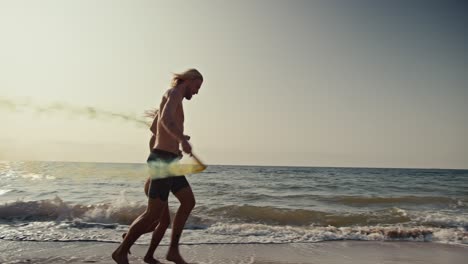 A-blond-guy-holds-his-blonde-girlfriend-in-an-orange-swimsuit-and-they-run-along-the-orange-brown-seashore-holding-smoky-fireworks-in-their-hands-that-leave-behind-green-and-yellow-colors-on-the-seashore-in-the-morning