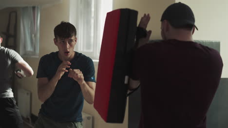 man trainer holds wrestling equipment while focused guy kicks target. working out with expert in basement of sports club. gaining energy from doing exercises