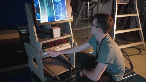person kneeling and adjusting settings on video equipment in tv studio. monitor shows broadcast setup, timer on control panel displays time. studio lighting and cables visible in background