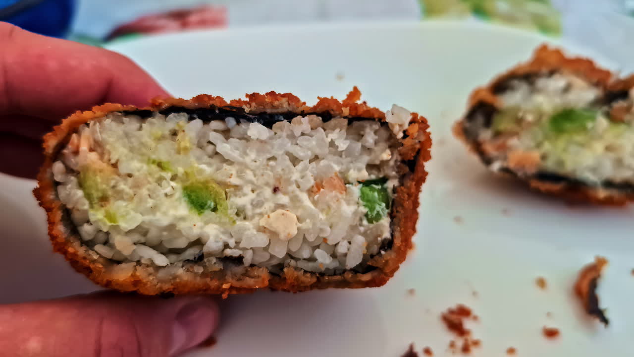 Close-up view of hand holding deep-fried sushi roll with rice, vegetables and seafood filling