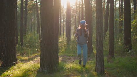 young woman walking through serene forest looking around, holding backpack straps as soft evening sunlight creates a gentle glow around her, tall trees and lush greenery frame her path