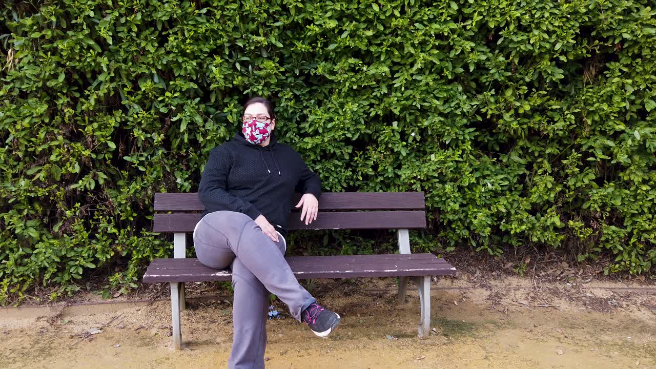 얼굴 마스크를 쓴 여성은 전염병으로 인해 공원 벤치에 앉아 주변을 둘러보고 카메라를 바라보고 있습니다.
