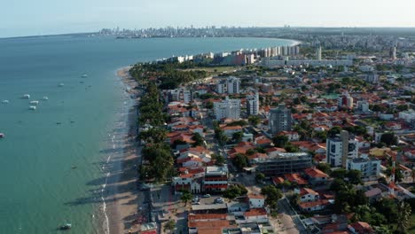 Incline-Hacia-Arriba-Un-Dron-Aéreo-En-Ascenso-Toma-Extrema-De-La-Playa-Tropical-Bessa-En-La-Ciudad-De-Joao-Pessoa,-Paraiba,-Brasil-Con-Gente-Disfrutando-Del-Océano,-Botes-Turísticos-Y-Un-Gran-Paisaje-Urbano-En-El-Fondo