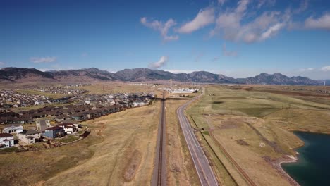 Following-the-train-tracks-and-road-in-the-Rocky-Flats-towards-the-Rocky-Mountains,-aerial