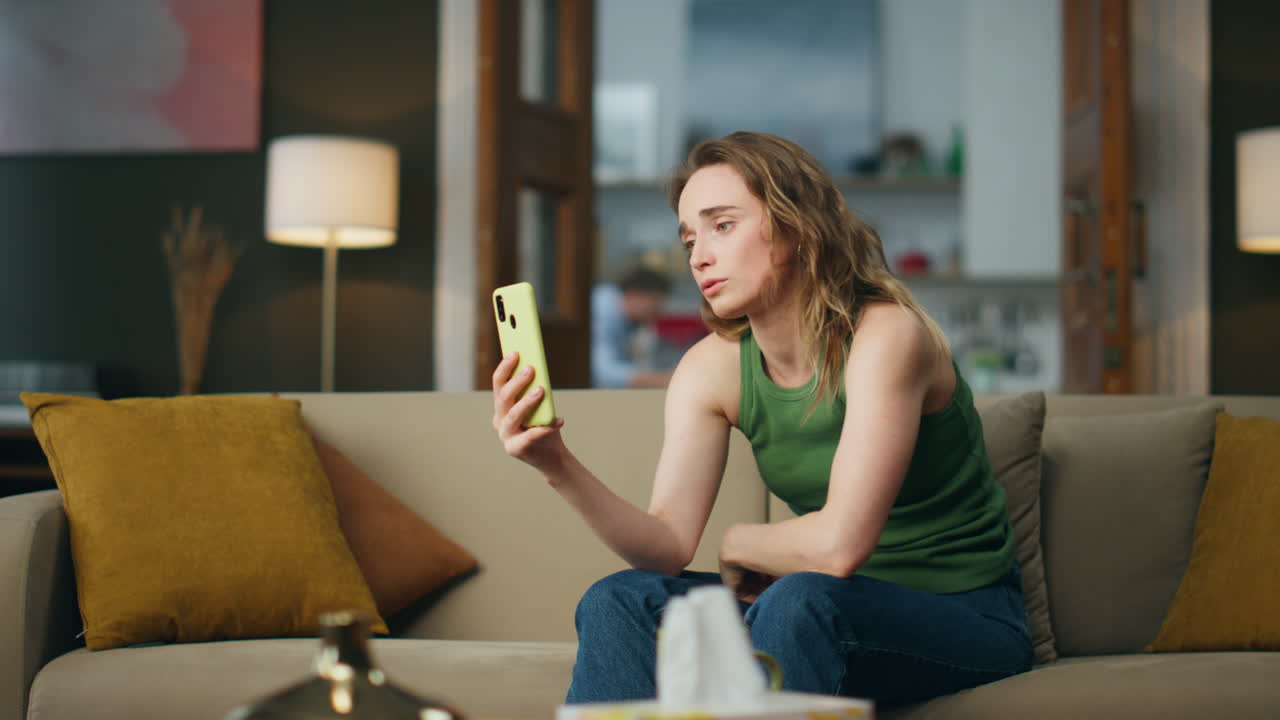 울고 있는 여자가 집 소파에서 스마트폰으로 얘기하고 있다. 불행한 소녀의 드폰 영상 통화