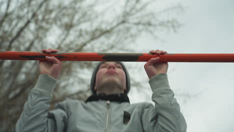 close-up of a boy working out on pull-up bars in an outdoor gym area, after successfully completing his third pull-up, he struggles to continue, with a bare tree in the background