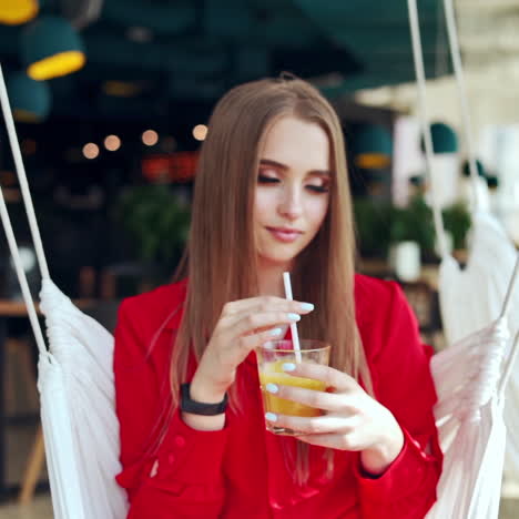 gorgeous woman drinking juice. beautiful young lady in red dress sitting on a swing and drinking cold beverage. attractive model relaxing in a cafe.