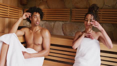video de una pareja relajada, feliz y diversa con toallas sentada y hablando en la sala de sauna en un spa de salud