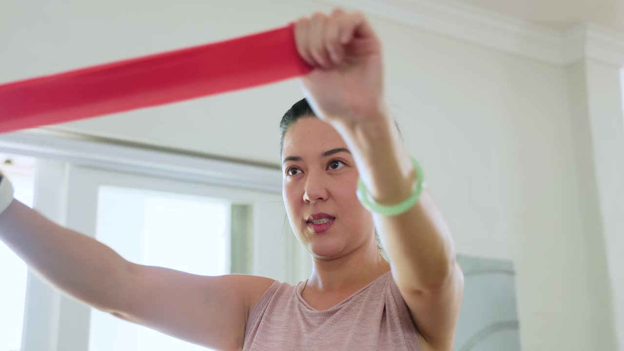 Donna adulta asiatica che solleva le braccia, allungando una fascia di resistenza rossa per un allenamento delle spalle vicino a una finestra.