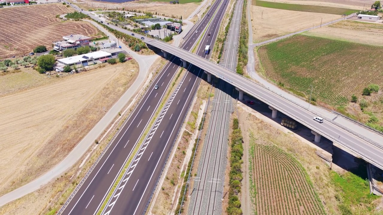 Riprese aeree che mostrano auto e camion che guidano lungo un'autostrada a più corsie accanto a una linea ferroviaria, con un moderno cavalcavia che le attraversa entrambe, circondati da campi agricoli in una giornata di sole