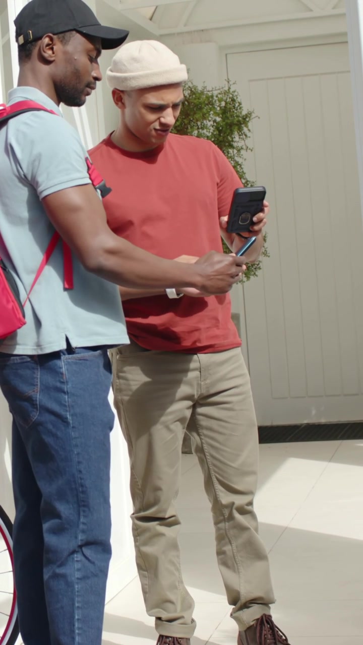 Vertical video: Coming courier with backpack showing phone as man tapping phones at door verifying
