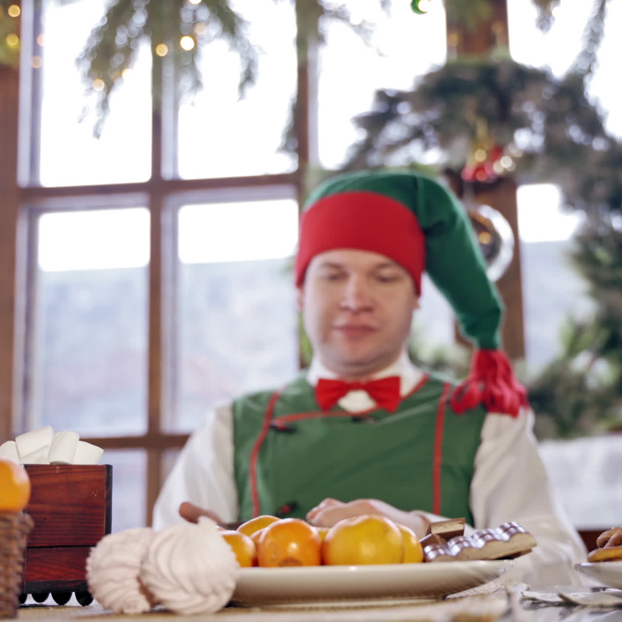 Hungry elf is impatiently tapping his feet at the Christmas table. Male elf in green costume with hood is sitting at festive table in front of the oranges. New Year and Christmas concept.