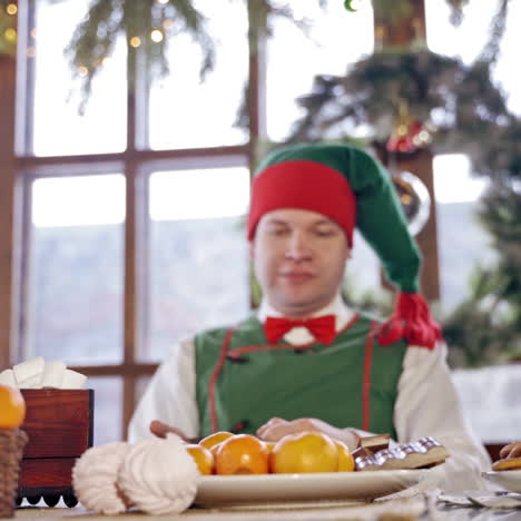 hungry elf is impatiently tapping his feet at the christmas table. male elf in green costume with hood is sitting at festive table in front of the oranges. new year and christmas concept.