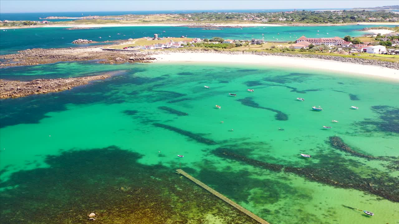 Drone footage over incredibly clear turquoise sea in bay with boats at anchor with shadows on sea bed in summer sun in north west Guernsey golden sandy beach and Napoleonic Martello tower .