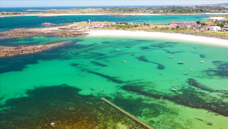 drone footage over incredibly clear turquoise sea in bay with boats at anchor with shadows on sea bed in summer sun in north west guernsey golden sandy beach and napoleonic martello tower .