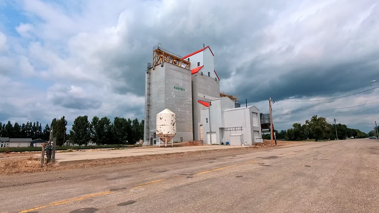 Hartney, MB Canada - July 19, 2024: The imposing Hartney grain elevator stands tall against a dramatic sky. White clouds gather, hinting at rain, while the road stretches towards the horizon.