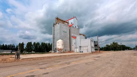 hartney, mb canada - july 19, 2024: the imposing hartney grain elevator stands tall against a dramatic sky. white clouds gather, hinting at rain, while the road stretches towards the horizon.