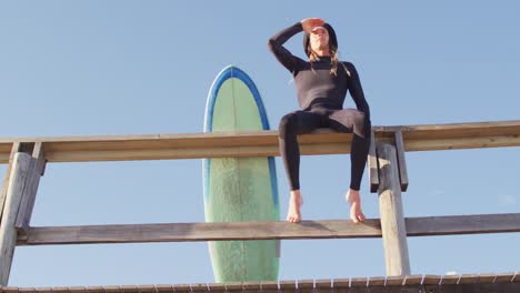 video of caucasian man with dreadlocks sitting on beach promenade with surfboard looking out to sea