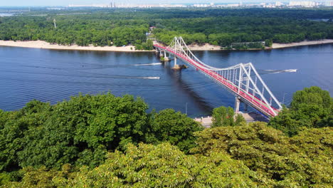 Volando-Sobre-árboles-Verdes-Para-Revelar-El-Puente-Peatonal-Del-Parque-Sobre-El-Río-Dnipro-En-El-Centro-De-Kyiv,-Que-Conduce-A-La-Isla-De-Trukhaniv