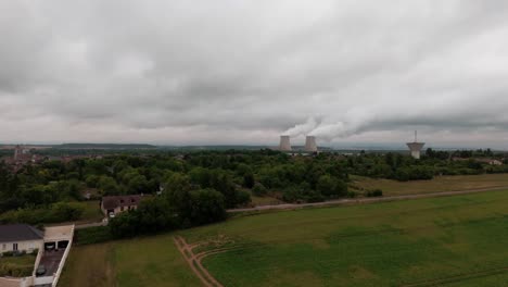 wide aerial drone shot of the french countryside with a nuclear power plant on the horizon. cooling towers release steam into the cloudy sky above a green landscape