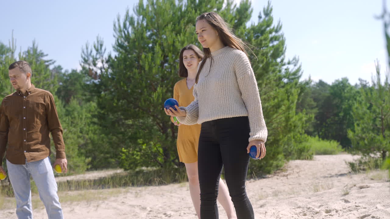 Side view of caucasian young woman throwing a petanque ball on the beach on a sunny day while her friends wait their turns